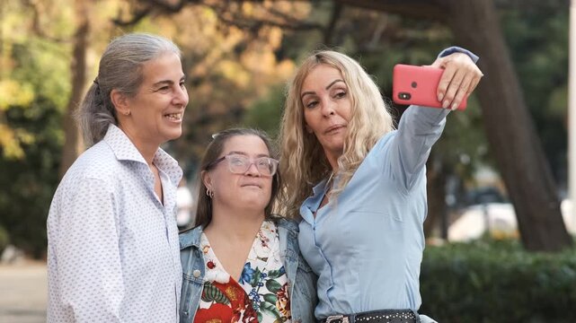 Slow motion of happy mature women and female friend with Down syndrome taking selfie with smartphone at park against blurred background during daytime