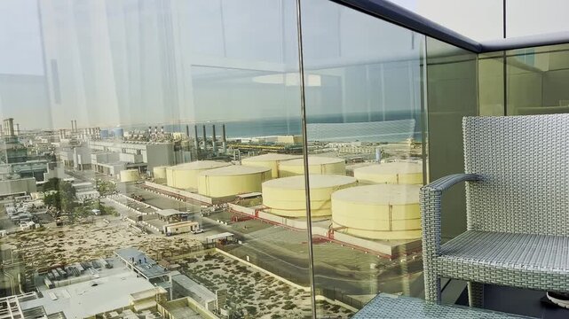 High vantage terrace scene overlooking coastal industrial complex with rows of pale storage tanks, pipes and chimney stacks, factory blocks, and calm sea beyond, framed by woven chair and glass