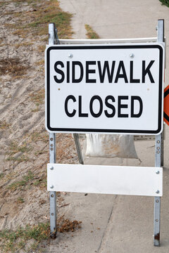White sidewalk closed sign with bold black text mounted on a metal frame placed near a sandy patch in San Diego, California