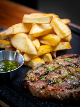 Close-up of a juicy grilled pork bondiola served with golden fried potatoes and a fresh herb sauce on a dark stone platter. The meat features pronounced grill marks, a browned crust, and a glossy herb