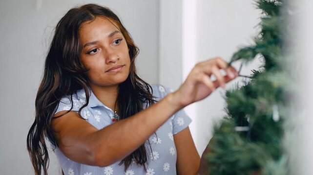 Beautiful young latina woman joyfully arranging lights on an artificial christmas tree, preparing for the holiday season at home