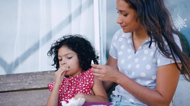 Charming little girl with curly hair eating a snack with her loving mother. They are sitting together on the porch steps, enjoying a moment