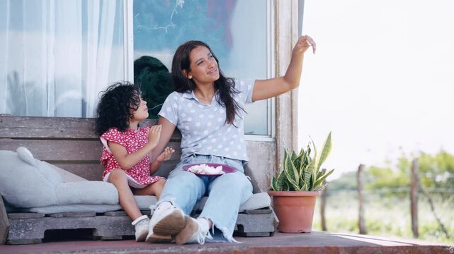 Beautiful young mother spending quality time with her adorable little girl on their porch. They are sitting, talking, and enjoying a snack