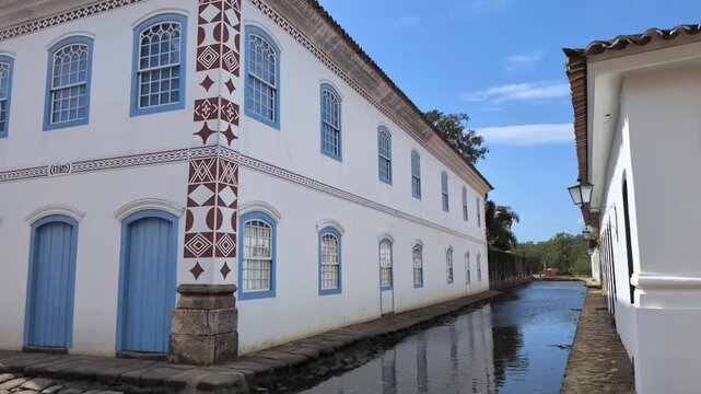 Static slow motion shot of historic colonial mansion in Paraty, Brazil, featuring masonic geometric wall details next to a flooded canal under a blue sky.
