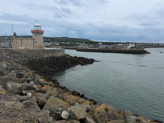 Historic Howth Harbour lighthouse stands beside a rocky breakwater and calm coastal water beneath an overcast sky in County Dublin, Ireland. © Ryan