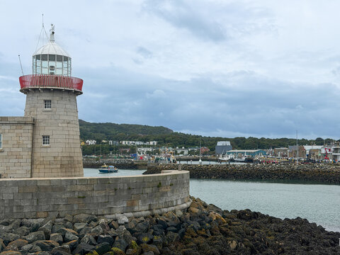Historic stone lighthouse overlooks Howth Harbour, with fishing boats, rocky breakwaters and the coastal village beneath a cloudy Irish sky.