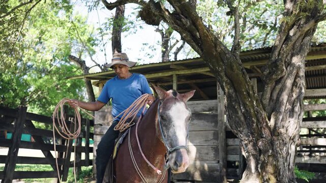 Latin man wearing a traditional hat, sitting on his beautiful brown and white horse, and holding a rope on a sunny day at the ranch