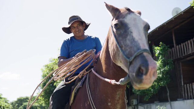 Low angle view of a smiling Latin stableman sitting on his horse. The man is holding a lariat and looking at the camera outdoors