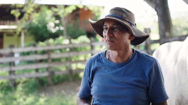 Hispanic farm worker with a hat standing next to a white horse. The man is looking away with a thoughtful expression in a rural setting