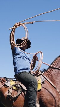 Latino man wearing a cowboy hat sitting on a brown horse. He is practicing his roping skills, skillfully twirling a lasso in the air