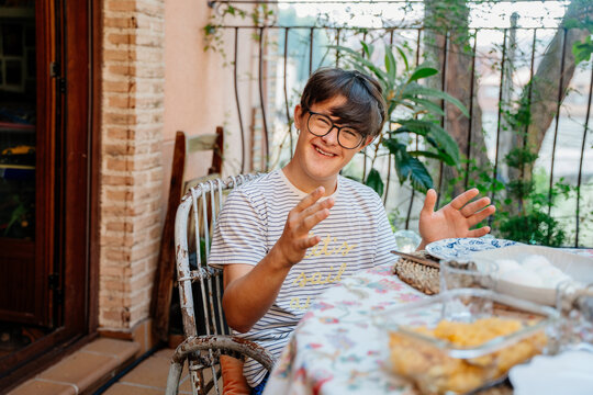Happy teenage boy with Down syndrome sitting at outdoor breakfast table on family terrace