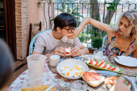 Caring mother wiping mouth of teenage son with Down syndrome during outdoor family breakfast