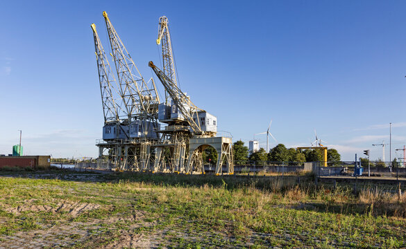 Antwerp, Belgium: Historic yellow dockland cranes standing as a monument in the port area, contrasting with modern wind turbines and industrial harbor infrastructure under a blue sky.