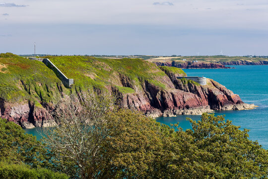Historic coastal defence walls built into rocky headland, lush gorse and bracken above red rock formations dropping into calm coastal water on sunny day