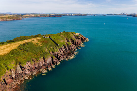 Rugged red sea cliffs topped with green gorse and stone fortifications, clear coastal waters below under bright summer sunlight, Welsh coastline
