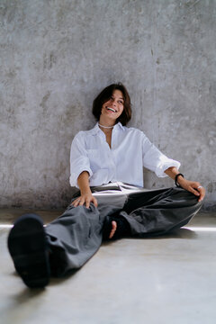 Fluid career hybrid lifestyle choices inspire cheerful Caucasian female nomad laugh joyfully while sitting cross legged on floor near rustic concrete wall backdrop inside studio room