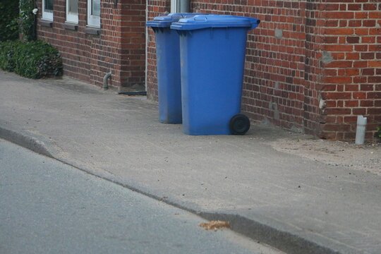 two blue colored garbage cans stand on the street at the house wall