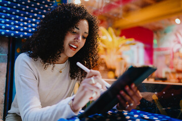 Happy young Spanish woman holding digital tablet and drawing with pencil tool in cozy cafe, cheerful female student e-learning online, modern technology lifestyle portrait, side view © BullRun