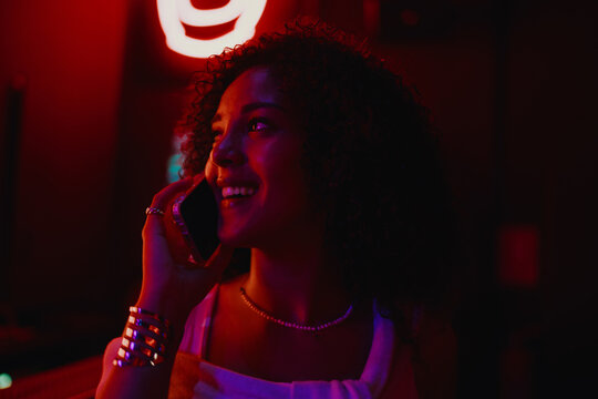 Close up of joyful young woman talking on mobile phone under red neon lights in night club, calling friends or scheduling meeting, active youth lifestyle and communication concept