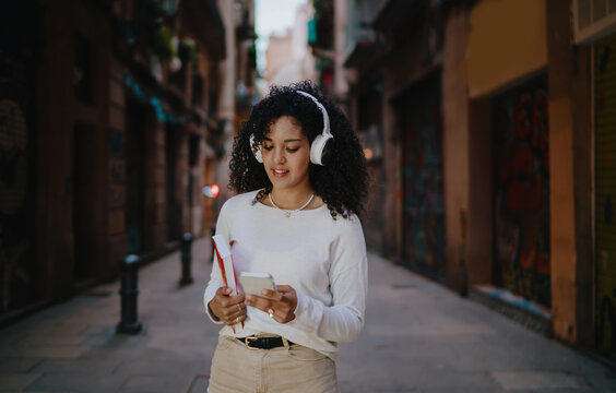 Smiling young woman in wireless headphones holding smartphone and red book while standing on cozy city street, female student listening to audio literature or music podcast portrait