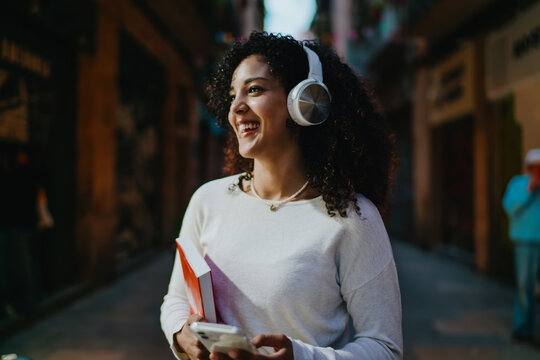Close up of smiling young woman in wireless headphones holding smartphone and book while walking on vintage city street, female student listening to music or online audio education