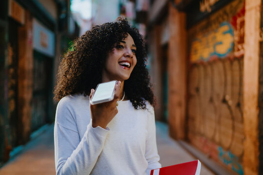 Cheerful young woman recording voice message on smartphone while walking on cozy street of European city, happy female tourist holding red book and enjoying modern communication