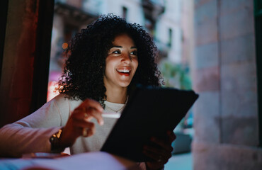 Cheerful young woman holding stylus pen and digital tablet laughing outdoors on evening city street, happy mixed race student enjoying online education process via smart modern tech gadget © BullRun