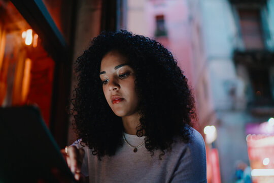 Focused young woman reading data on digital tablet screen standing on evening city street, creative freelance graphic designer working remotely utilizing smart portable device outdoors