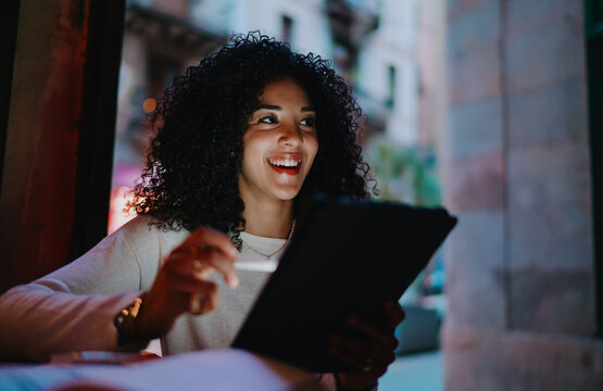 Cheerful young woman holding stylus pen and digital tablet laughing outdoors on evening city street, happy mixed race student enjoying online education process via smart modern tech gadget