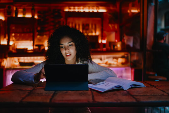 Serious young woman looking at digital tablet screen while sitting at cafe table with open book and smartphone, female freelance designer checking project details during remote work