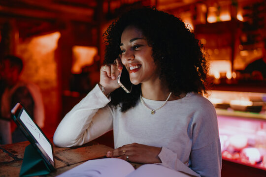 Smiling female freelancer talking on smartphone while analyzing business chart on digital tablet screen in cozy evening cafe, creative designer enjoying remote work and mobile communication