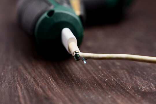 Damaged frayed power cord of an electric tool on a wooden surface. Broken white charger cable insulation with exposed wires.
