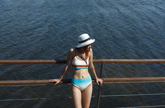 Young woman in a white sun hat and blue bikini standing on a wooden waterfront deck. Summer lifestyle scene focused on relaxation, travel and leisure by the water.