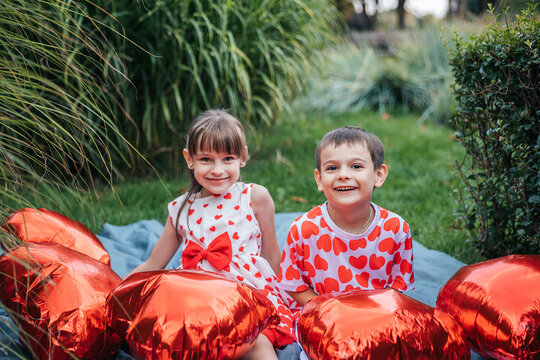 Two Children Celebrate Summer in a Garden Surrounded by Red Heart-Shaped Pillows on a Sunny Day