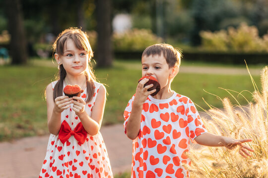 Two Children Enjoying Cupcakes in a Park During Summer While Wearing Matching Heart-Themed Clothing for Valentine's Day