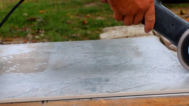 A skilled worker cuts a large ceramic tile using an angle grinder. Hands in work gloves hold the power tool tightly. Sparks fly in all directions.
