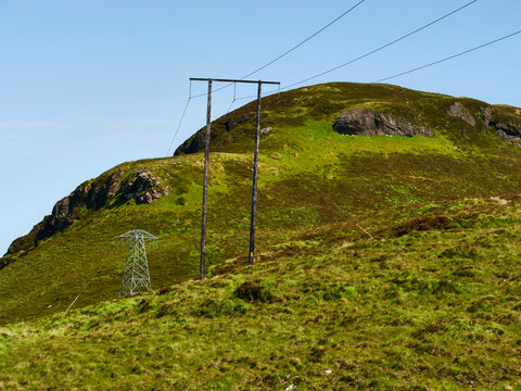 Two weathered wooden utility poles stand tall on a grassy hill. Wires stretch across clear blue sky. Green moss covers rounded hillside beneath. Scene evokes quiet solitude and rural resilience