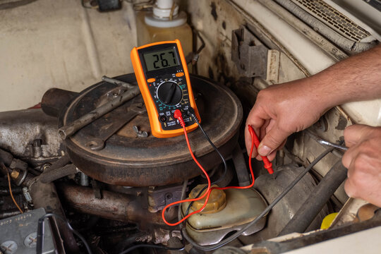 In a well-used garage, hands skillfully use a multimeter to check an engine component. The vintage car's parts reflect years of dedication and care