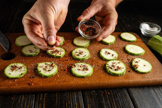 Hands gently sprinkle spices over freshly sliced zucchini on a wooden cutting board. The scene captures a delightful moment of preparing a healthy snack, showcasing the textures and colors