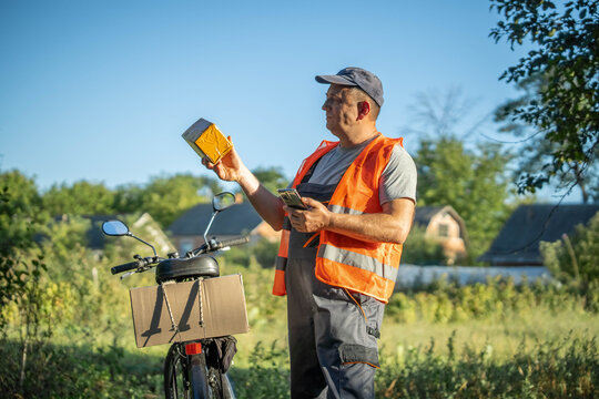 A delivery worker in an orange vest stands near his bicycle. He examines a package while surrounded by green fields and distant houses under a clear blue sky