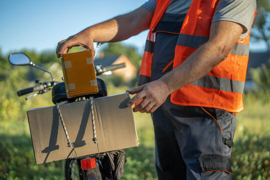 A delivery person wearing a bright vest secures packages on a bicycle in a lush, green area. The sun shines brightly, creating a warm atmosphere as they prepare for their next stop