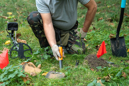 A treasure hunter kneels in a lush park, eagerly exploring the ground with a metal detector. Bright red flags mark where he has found intriguing artifacts, hinting at exciting discoveries