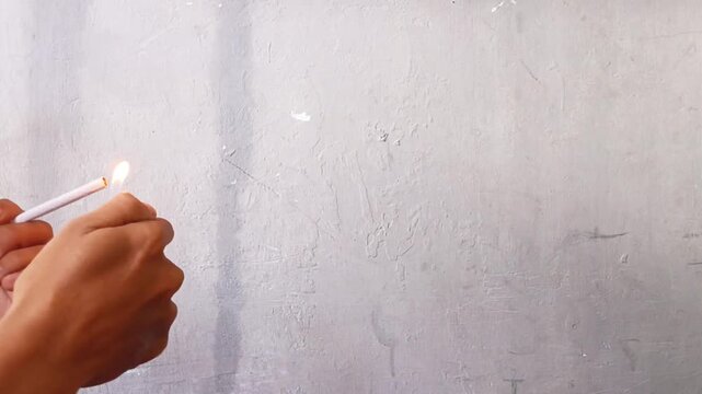 Hands lighting a cigarette with a burning match against a simple wall background, showing fire, smoke, ignition, and everyday smoking activity.
