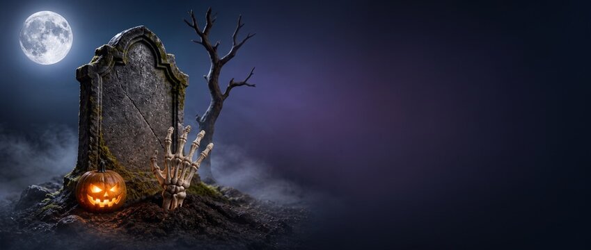 > Spooky Halloween graveyard background banner. Skeleton hand rising from the grave, tombstone, glowing pumpkin and full moon in the fog. Wide format with large copy space