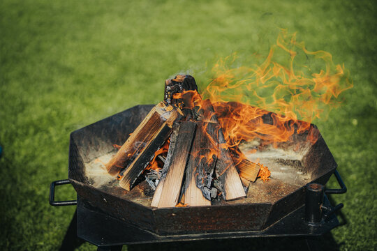 Firewood burning in a metal fire pit with flames rising against a green grass background, creating a warm outdoor atmosphere for gatherings and relaxation