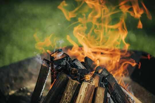 Close-up of burning firewood in a fire pit with vibrant flames and smoke rising against a blurred green grass background