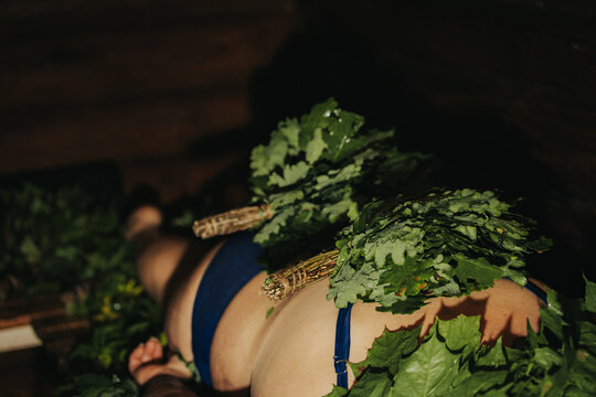 Person lying on a wooden surface covered with green leaves and branches, wearing blue swimwear, in a dimly lit indoor setting with wooden walls