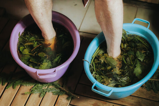 Bare feet submerged in two buckets filled with water and herbs, one purple and one blue, placed on wooden floor with natural elements around