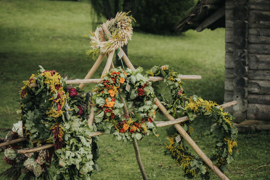 Colorful floral wreaths made of various flowers and greenery displayed on a wooden stand in a lush outdoor setting near a rustic wooden structure