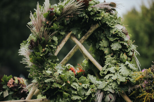 Decorative green wreath made of assorted leaves and flowers arranged on a wooden frame, showcasing natural elements in an outdoor setting